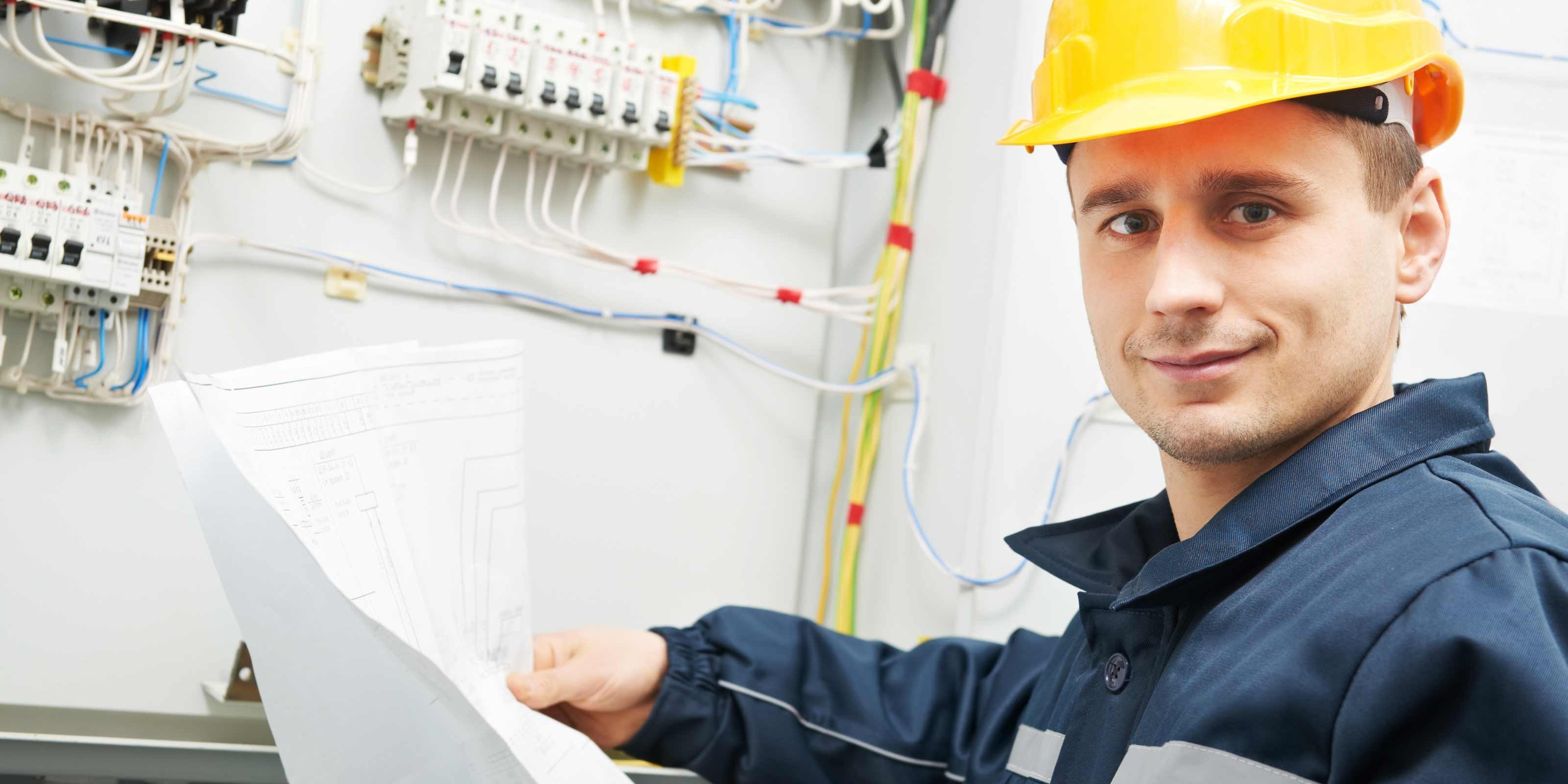 Ein Elektriker mit einem Bauhelm hält eine Zeichnung in der Hand vor einem Schaltschrank